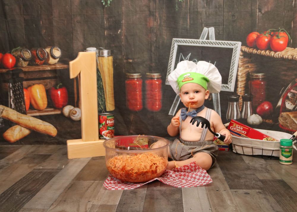 Baby in a chef hat with wooden number one and birthday cake in rustic kitchen setting.