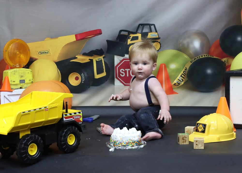 Baby surrounded by yellow toy construction trucks and balloons in studio setting.