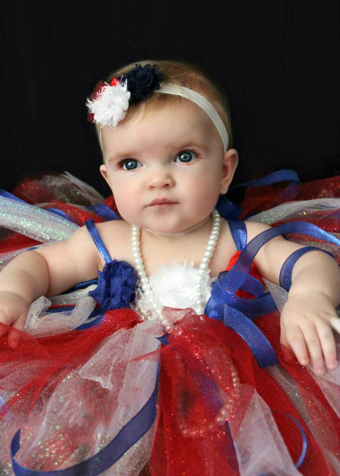 Baby wearing red white and blue tutu dress with pearls and hair accessories against black background.