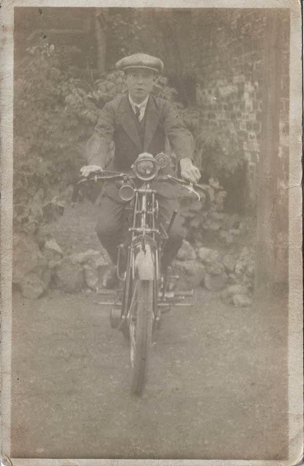 Vintage photo of person on early motorcycle wearing flat cap and suit against stone wall.