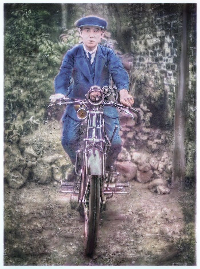 Colorized vintage photo shows motorcyclist in blue suit and cap posing with early motorcycle.