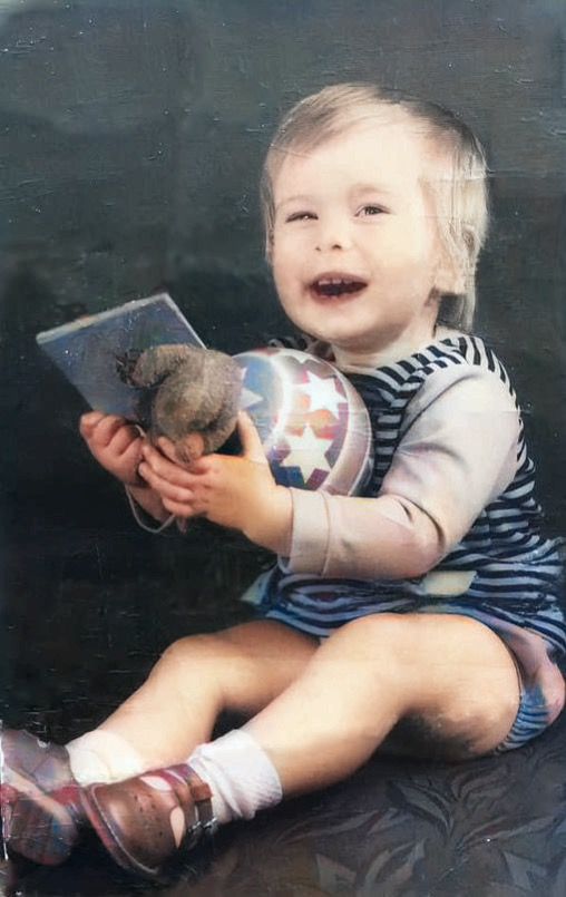 A toddler in a striped outfit smiles while holding a stuffed toy and photo in vintage photograph.