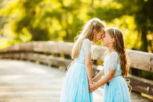 Two young children in matching light blue dresses share a special moment on a wooden bridge.