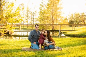 A family enjoys a picnic together in a sunny park with a wooden bridge in the background.