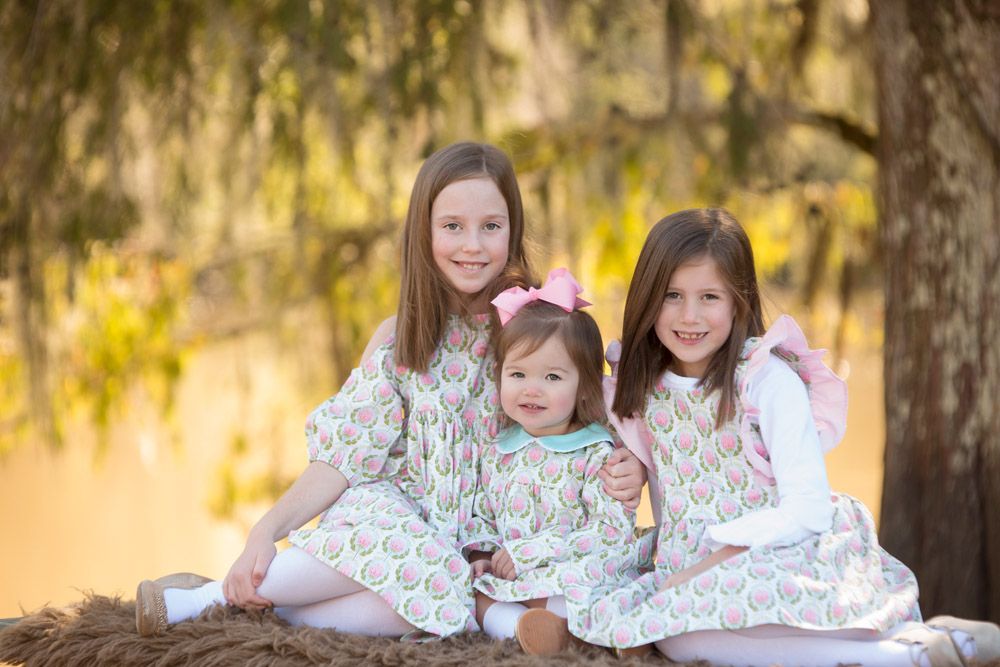 Three siblings in matching floral dresses sit together on a fallen tree during golden hour in nature.