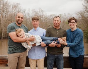 Four adults playfully hold and lift a smiling baby together outdoors on an autumn day.