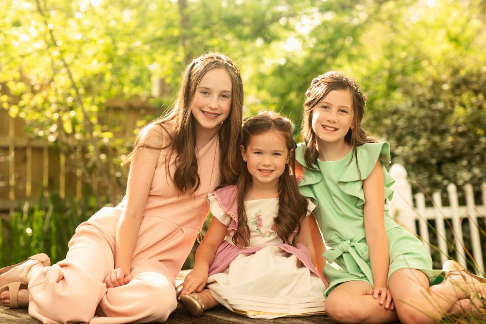 Three young children sit together on a bench in matching pastel dresses in a garden setting.