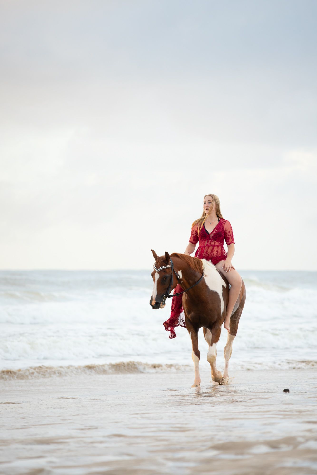 A girl in a red see through dress rides a chestnut pinto horse along a misty beach