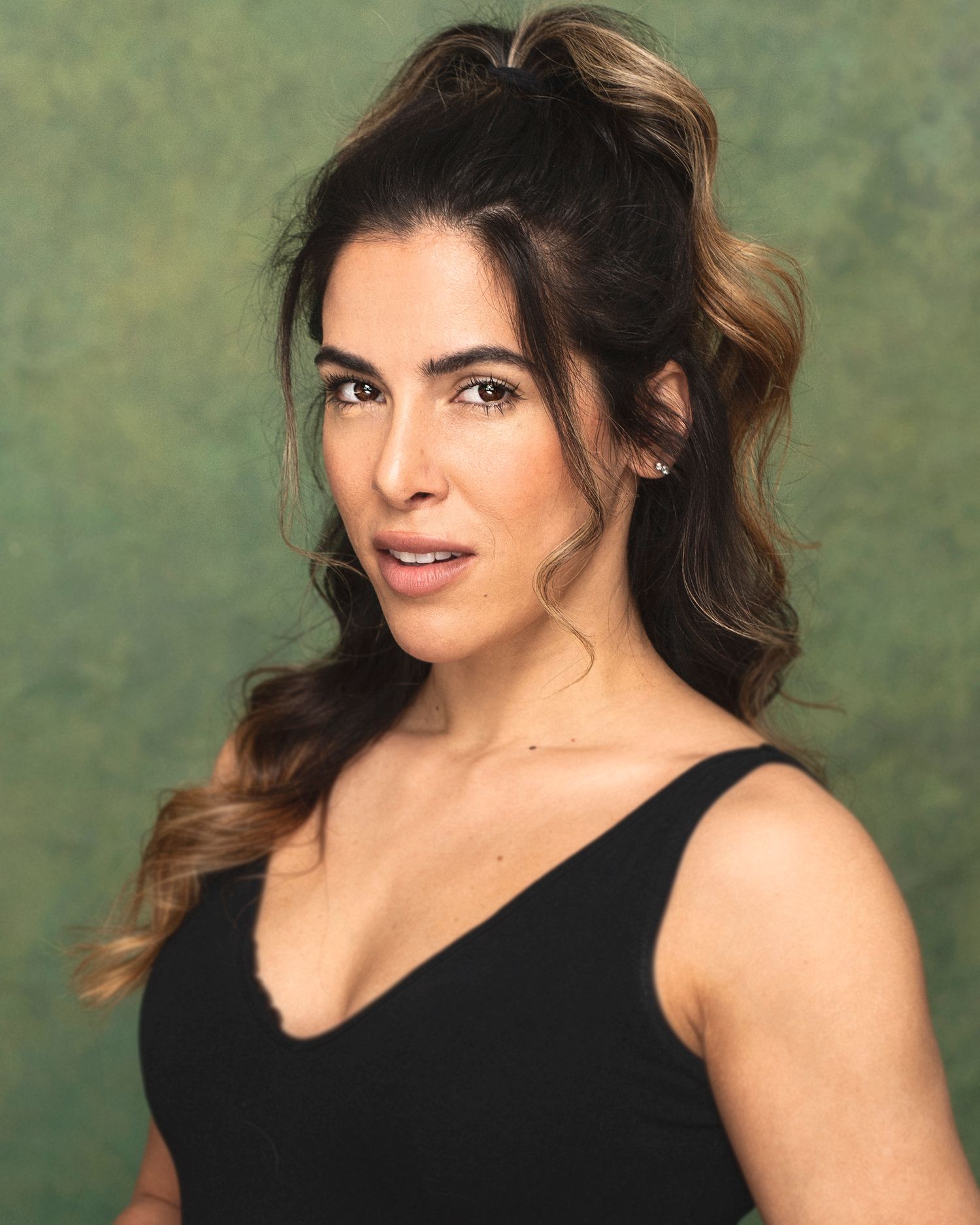Glamour headshot of a woman with wavy brown hair in a black tank top against a green background.