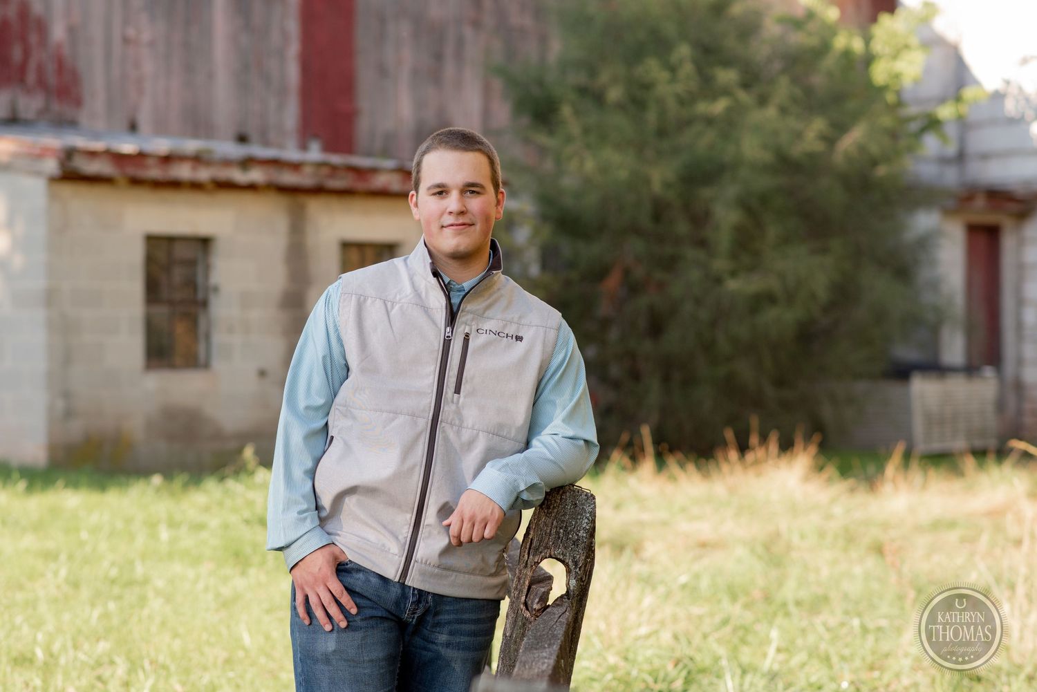 Person in light vest and denim jeans stands casually near rustic building in outdoor setting.