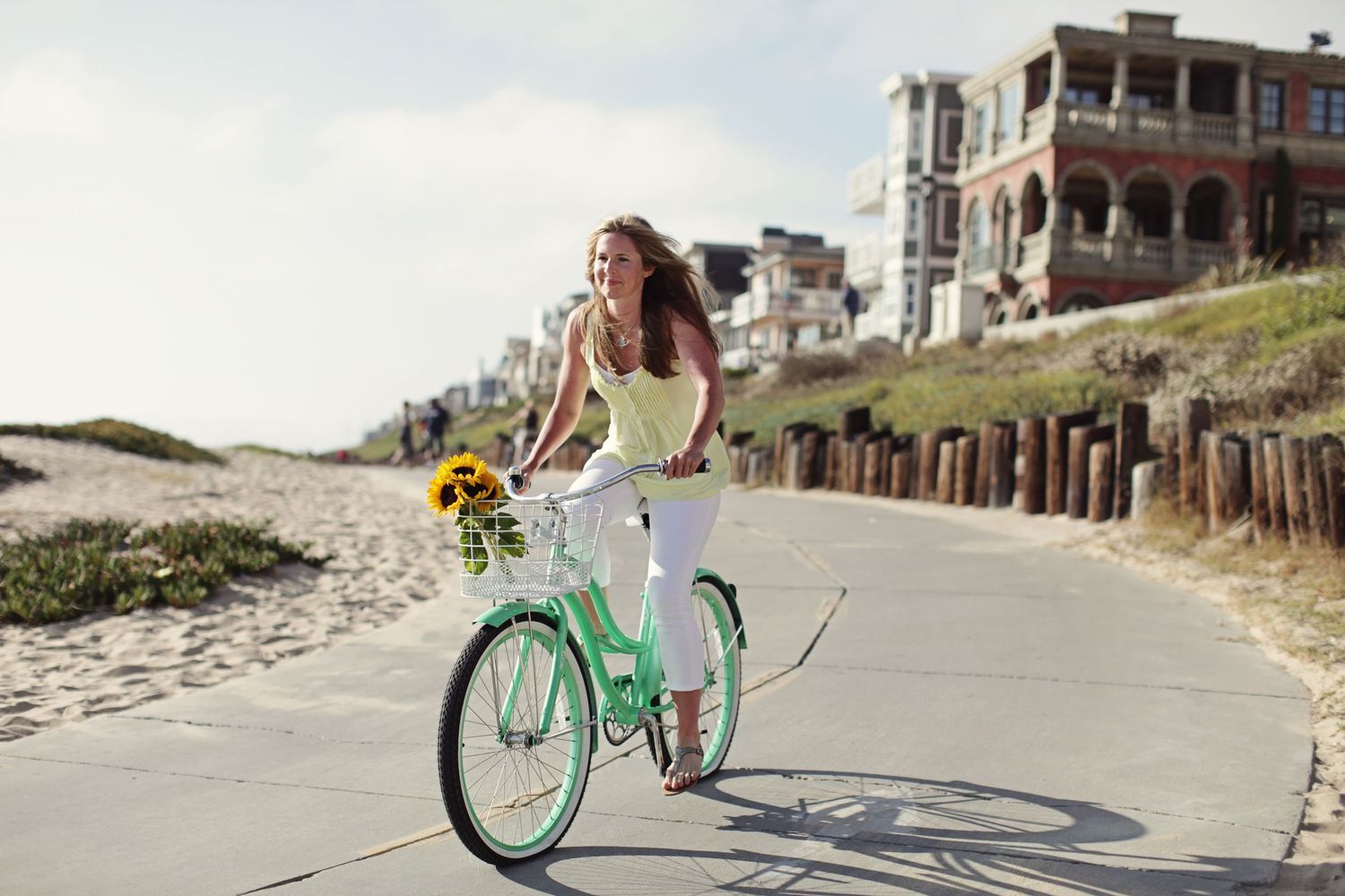 Woman riding mint green bicycle along coastal path with beachfront houses.