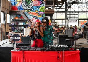 DJs perform behind red-draped tables at an indoor venue with colorful graffiti wall art in the background.