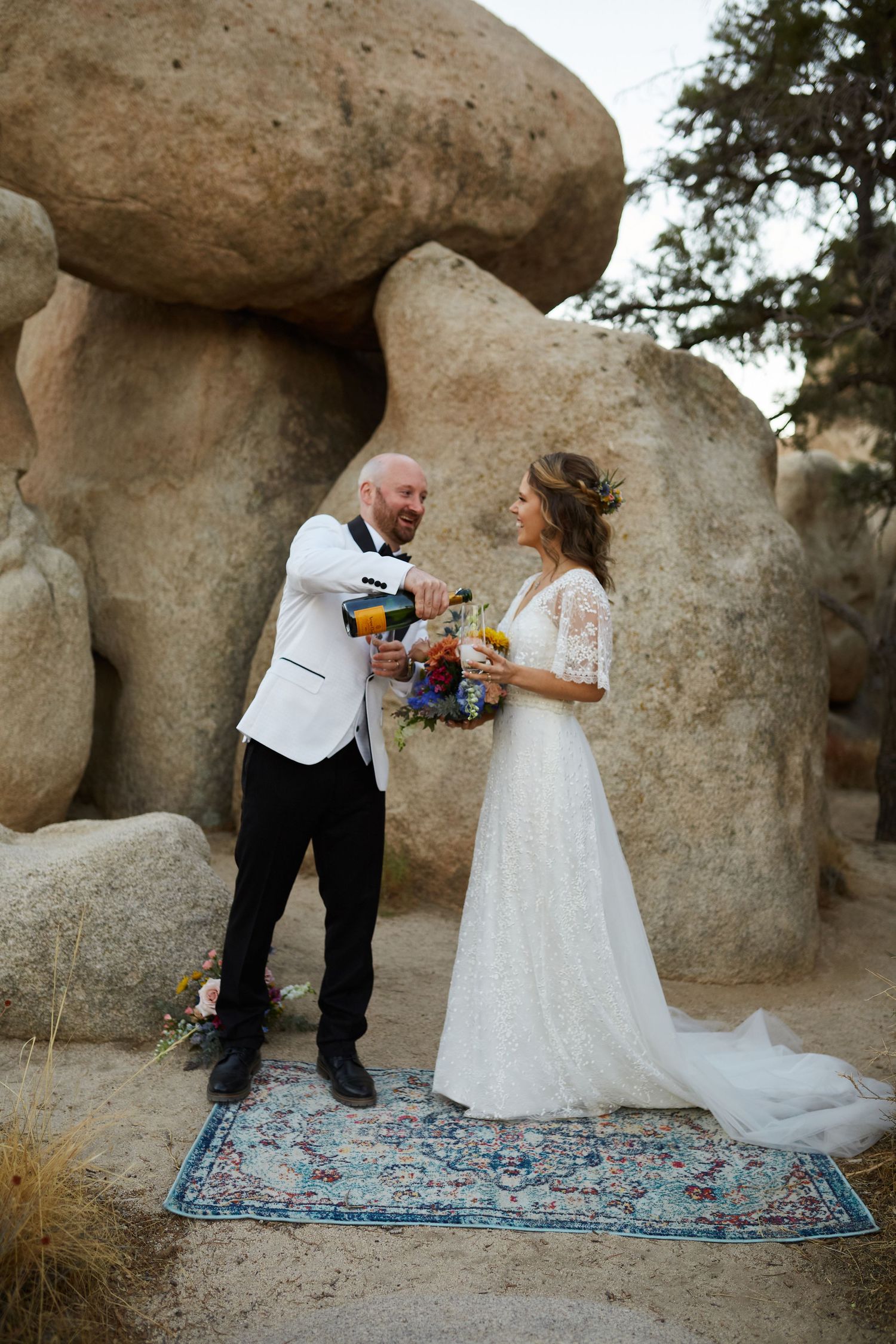 A couple poses on a decorative rug among large desert boulders in formal attire.