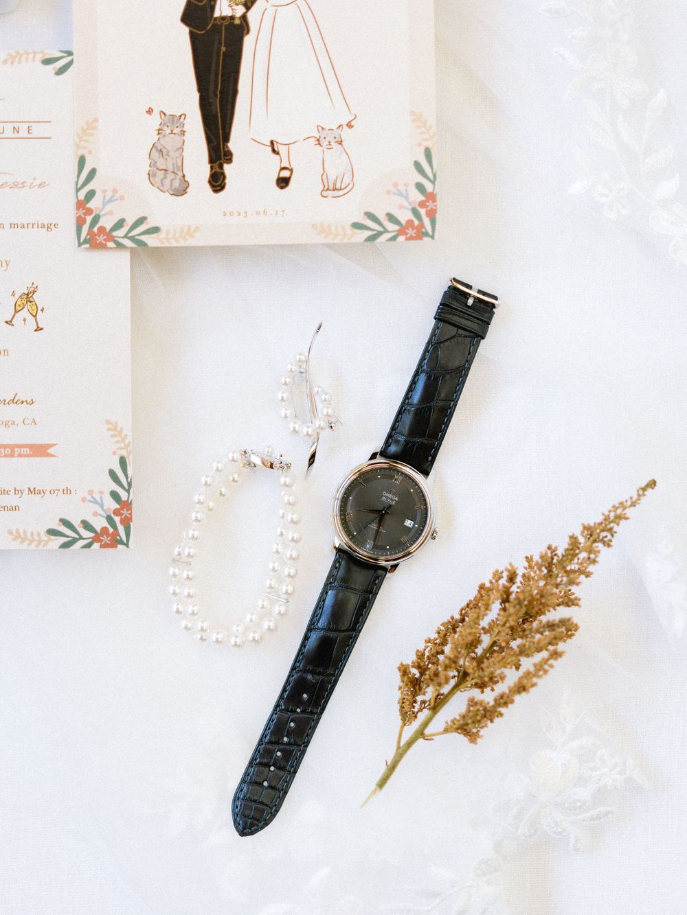 Wedding details including a watch, dried flowers and invitation with illustrated couple.