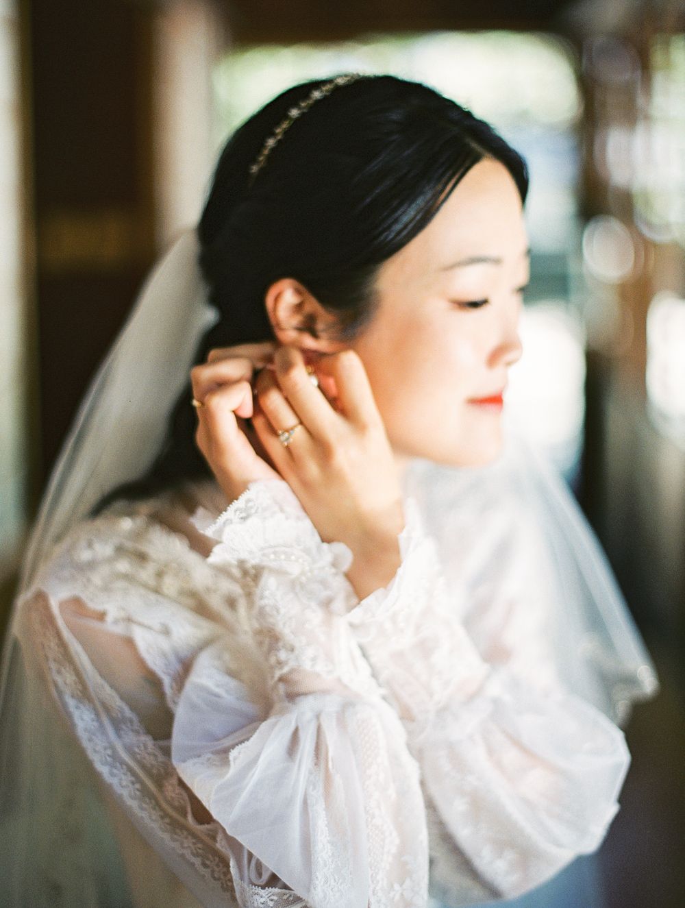 A bride wearing delicate lace gown and veil adjusts her earring in natural light.