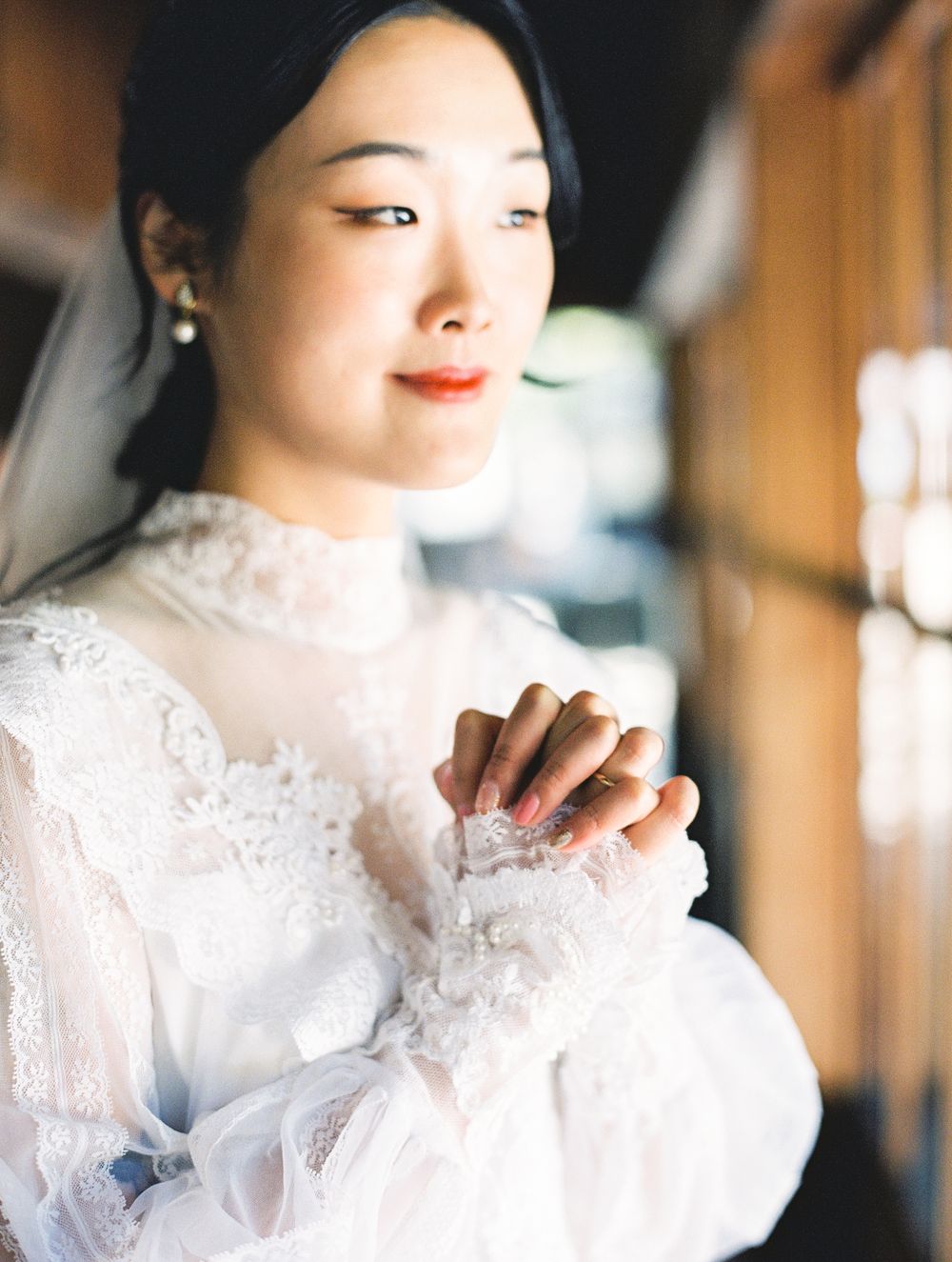 A bride in white lace wedding dress with high collar poses near window in soft light.
