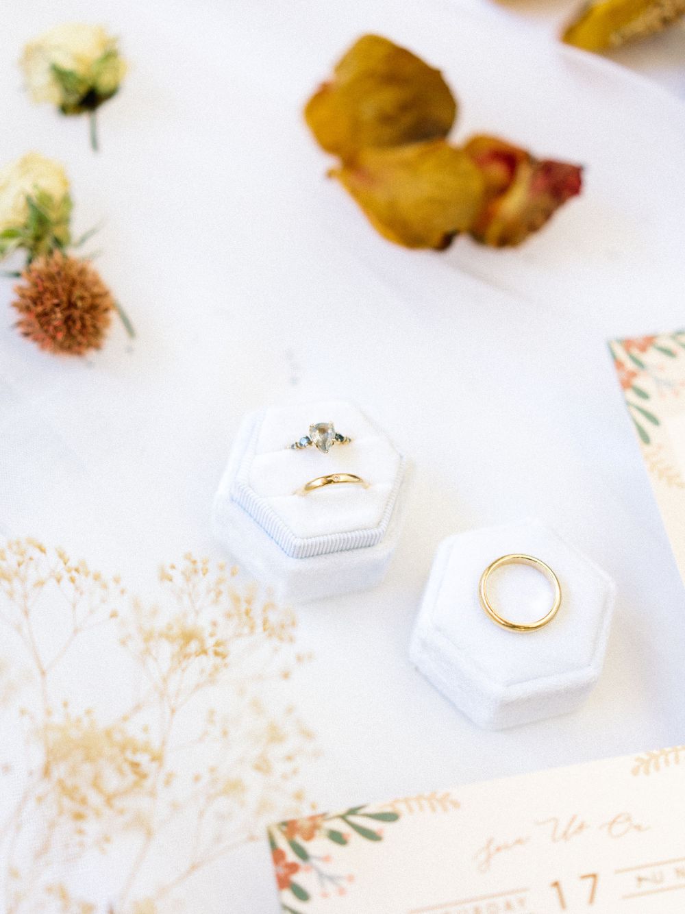Wedding rings in white hexagonal boxes with dried flowers and invitation details.