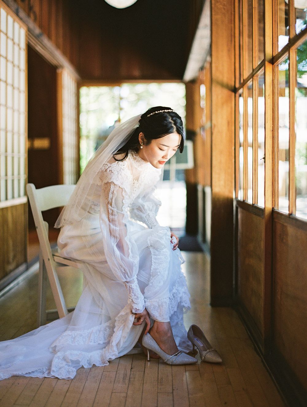 Bride in classic white dress adjusts her shoe in a wooden hallway.