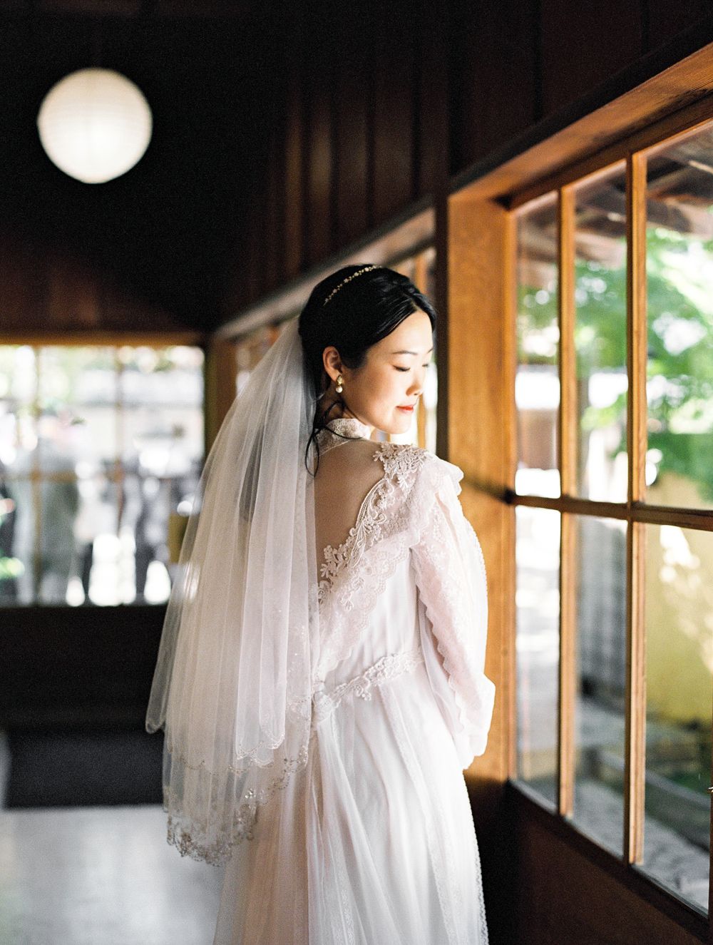 Bride in elegant long-sleeved lace gown and veil stands near wooden windows.