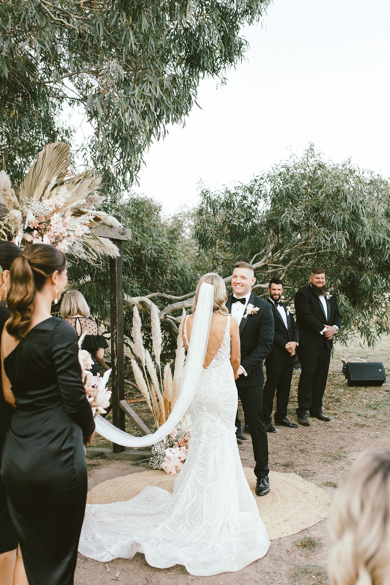 Wedding ceremony with elegant white lace gown and black tuxedos under willow trees.