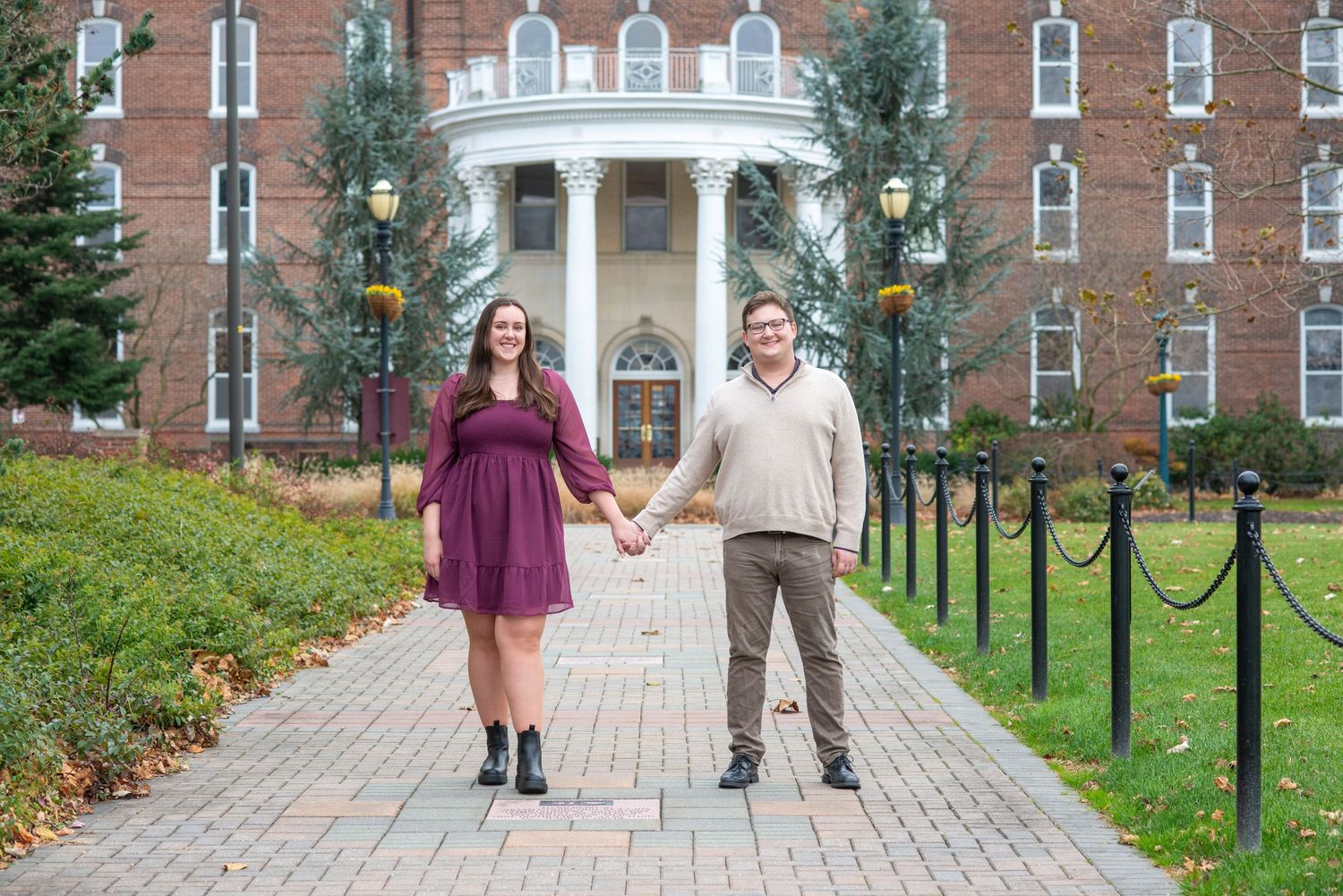 Two people hold hands while standing on a brick pathway leading to a grand collegiate building with white columns.