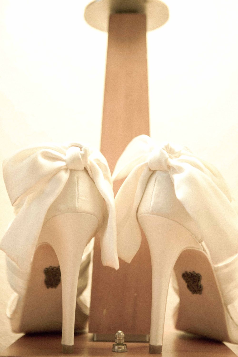 A pair of elegant white high heel bridal shoes with large bows on the back photographed from behind.