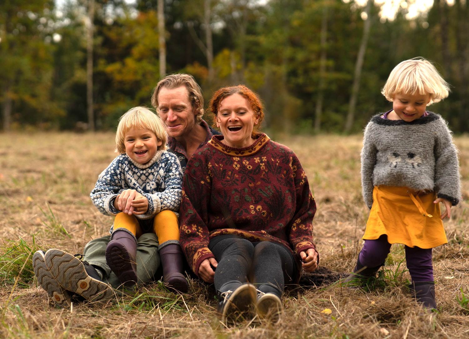 En familj med två små barn sitter på en gräsmassa och skrattar. Ett av barnen står upp.