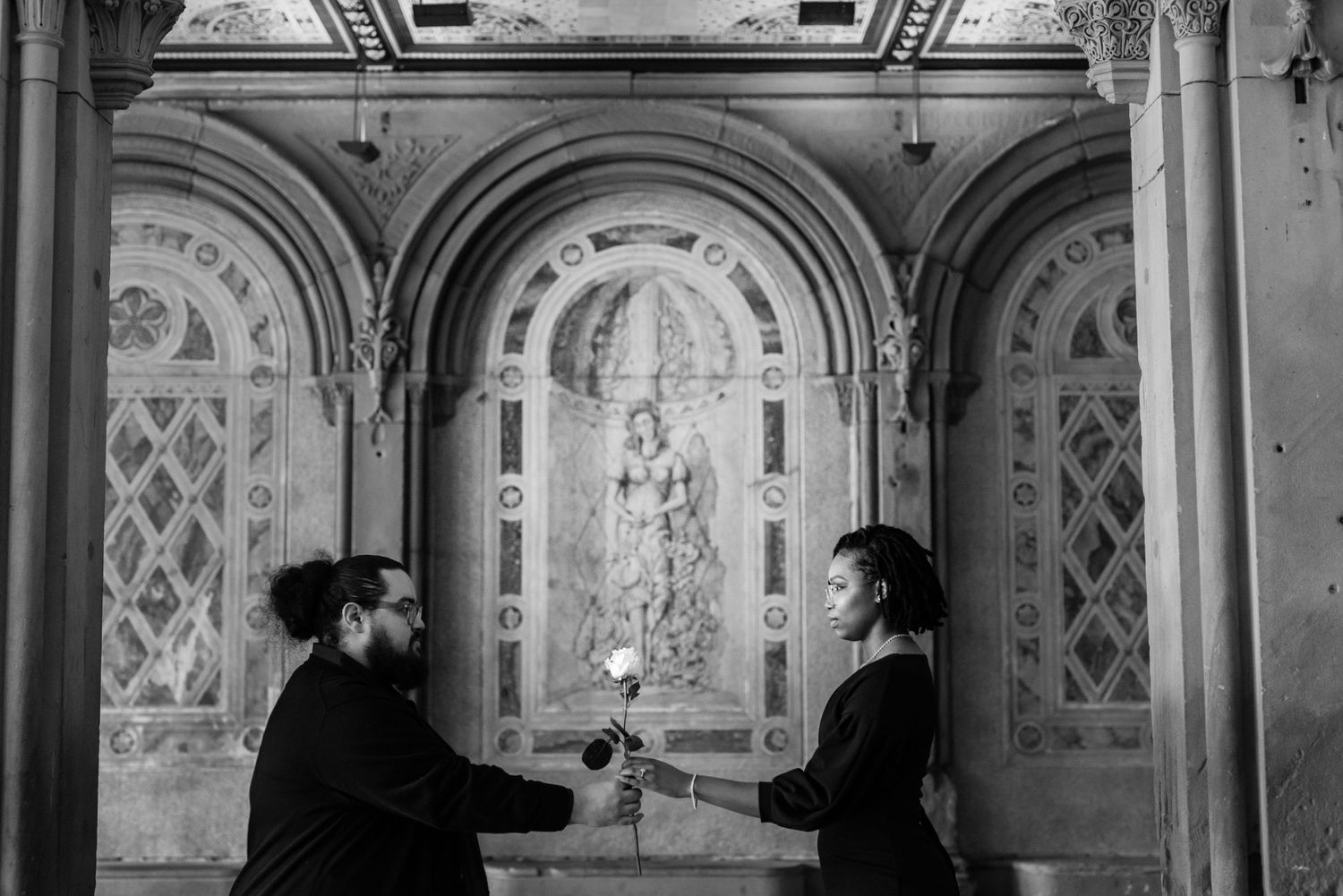 Two silhouettes exchange a flower in an ornate architectural setting with arched ceilings.