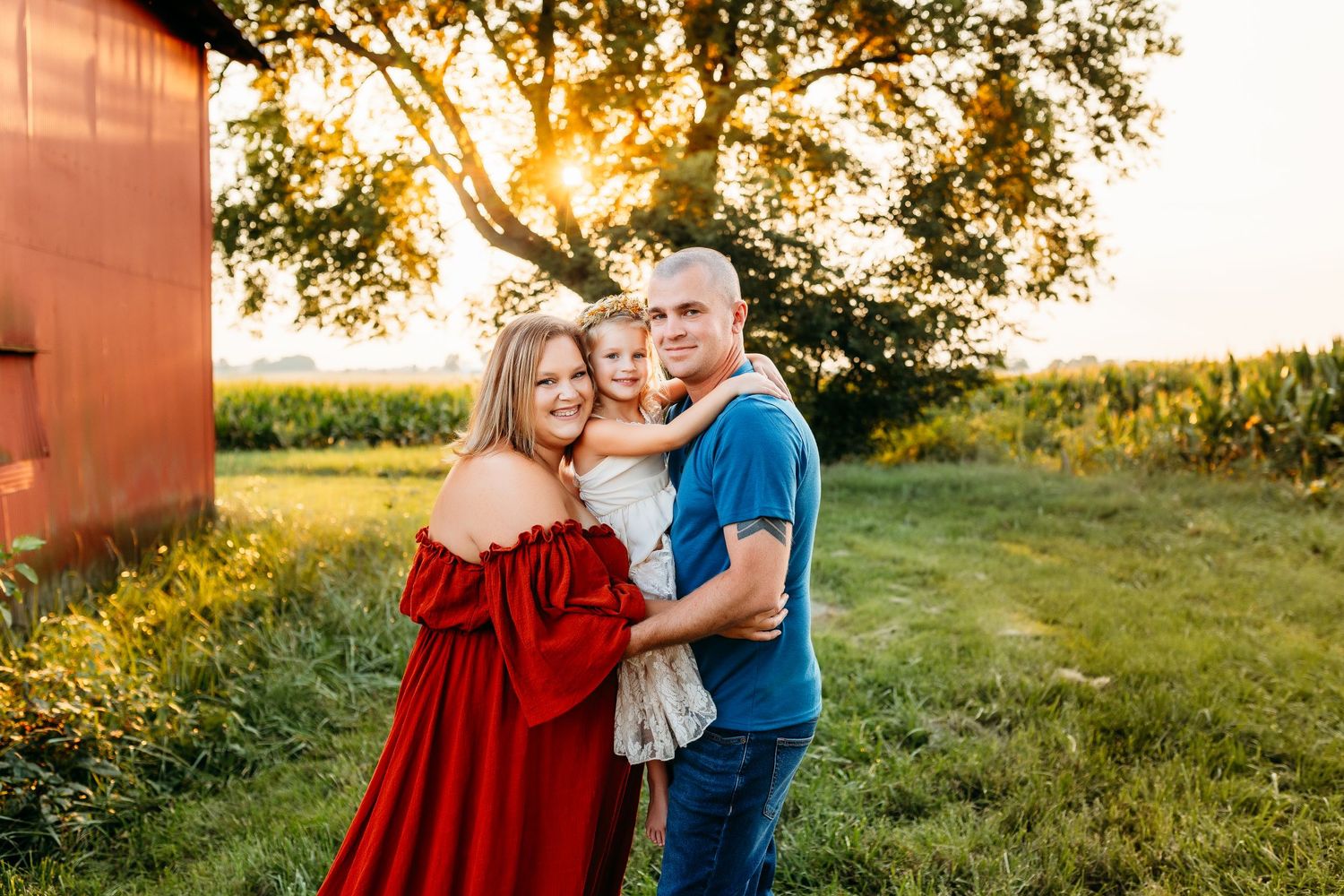 A couple poses together at sunset near a red barn in a rustic countryside setting.