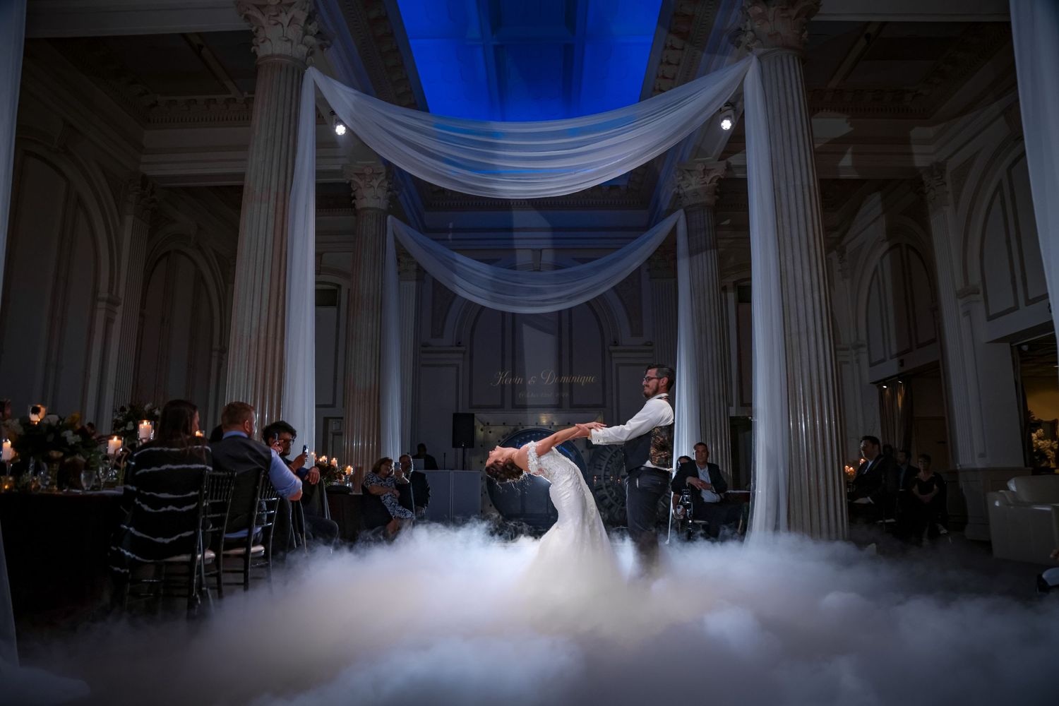 A couple shares their first dance on a misty dance floor under dramatic blue lighting and white draping in an elegant venue.