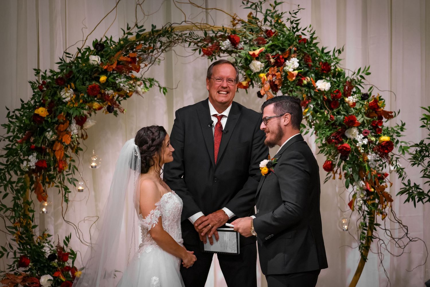 A wedding ceremony takes place under an elegant floral arch adorned with red and white roses and greenery.