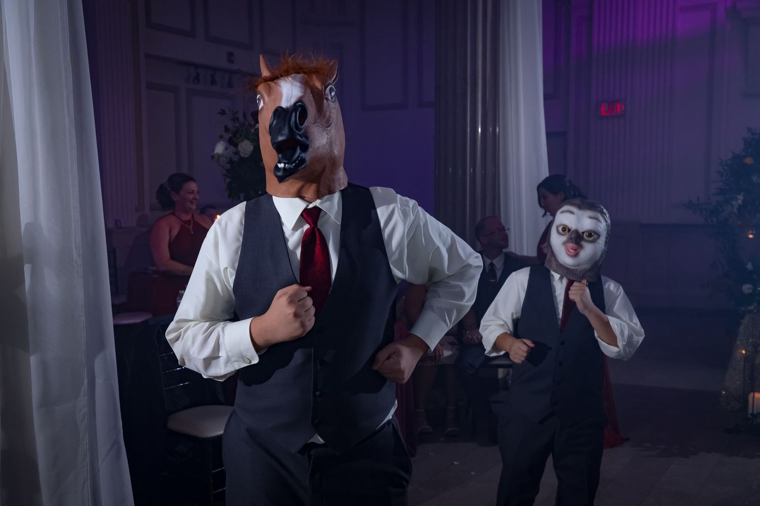 Two people in formal attire wearing horse masks dance at a reception in a dimly lit room with purple lighting.