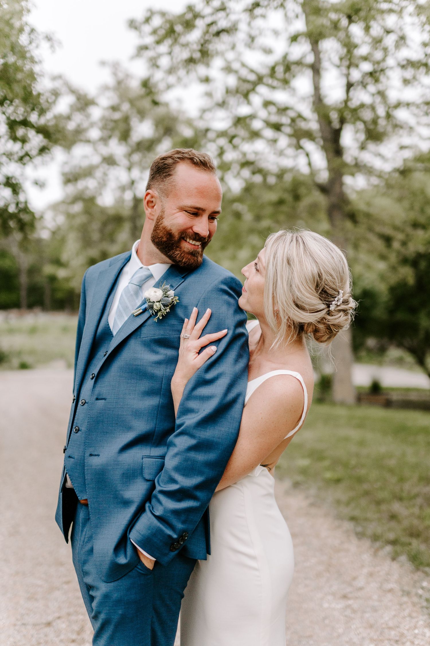 A couple shares a romantic moment on a gravel path surrounded by green trees.