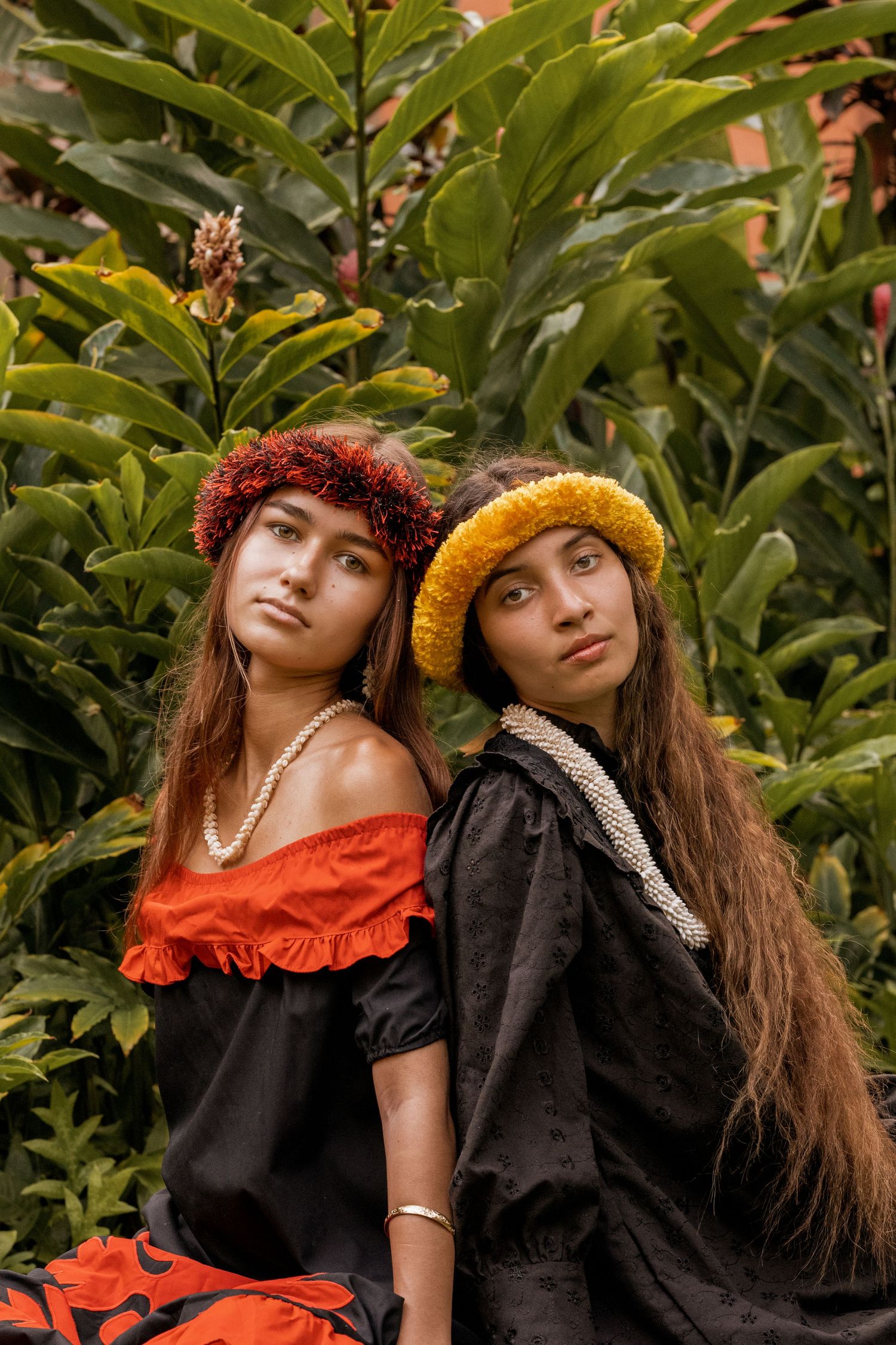 Two women wearing traditional Hawaiian headpieces pose against tropical foliage.