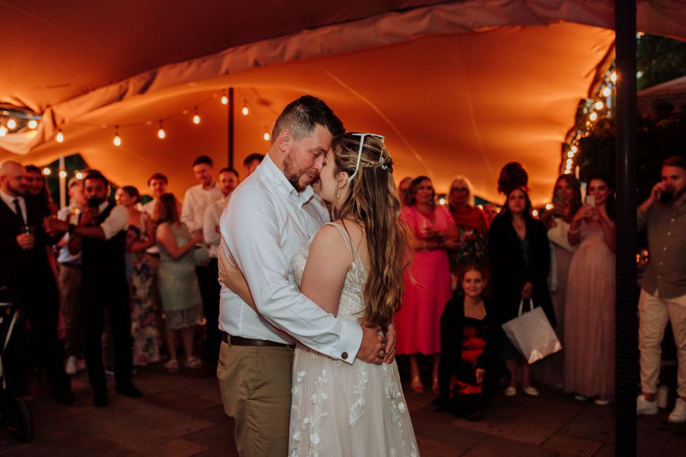 First dance under warm lighting in wedding reception tent with guests watching.