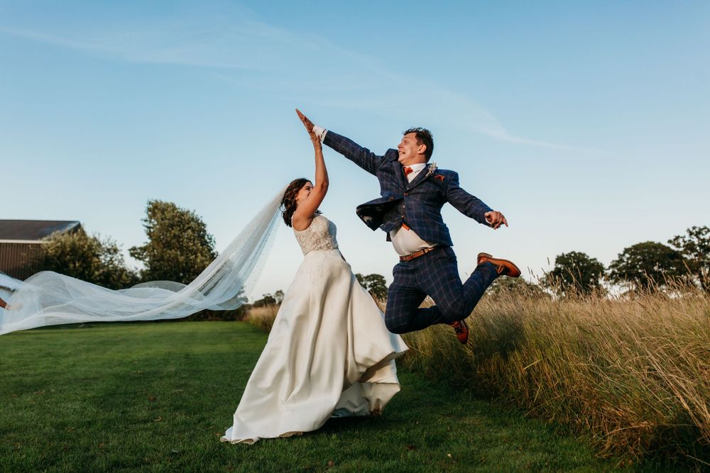 Joyful couple leaping together in formal attire against blue sky on their wedding day.