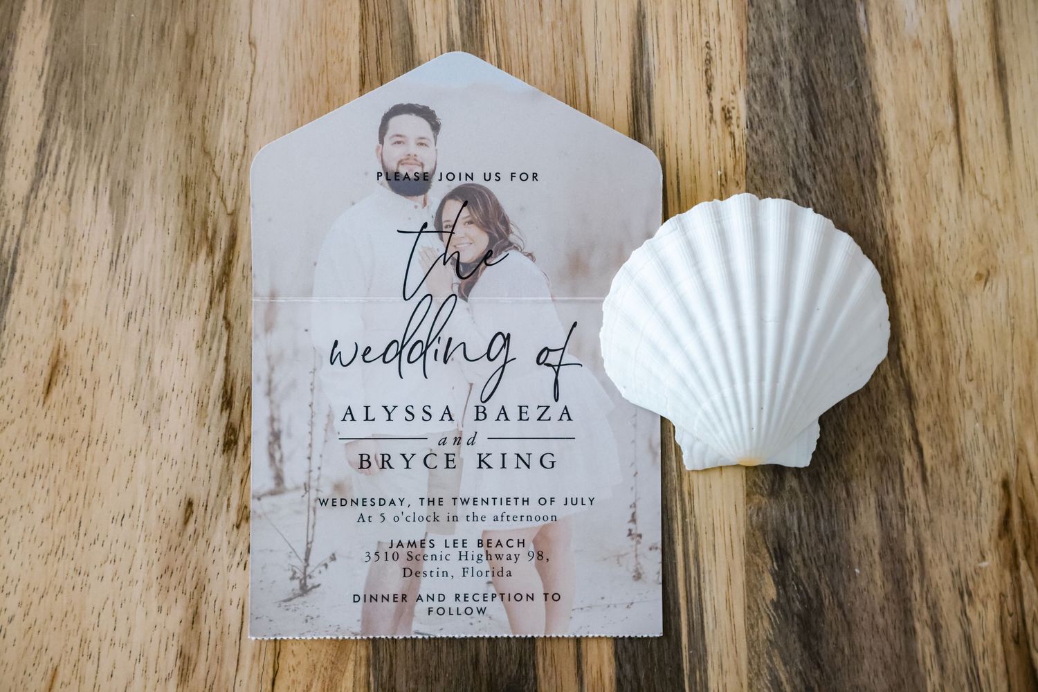 A wedding invitation with watercolor design displayed with a white seashell on wooden surface.