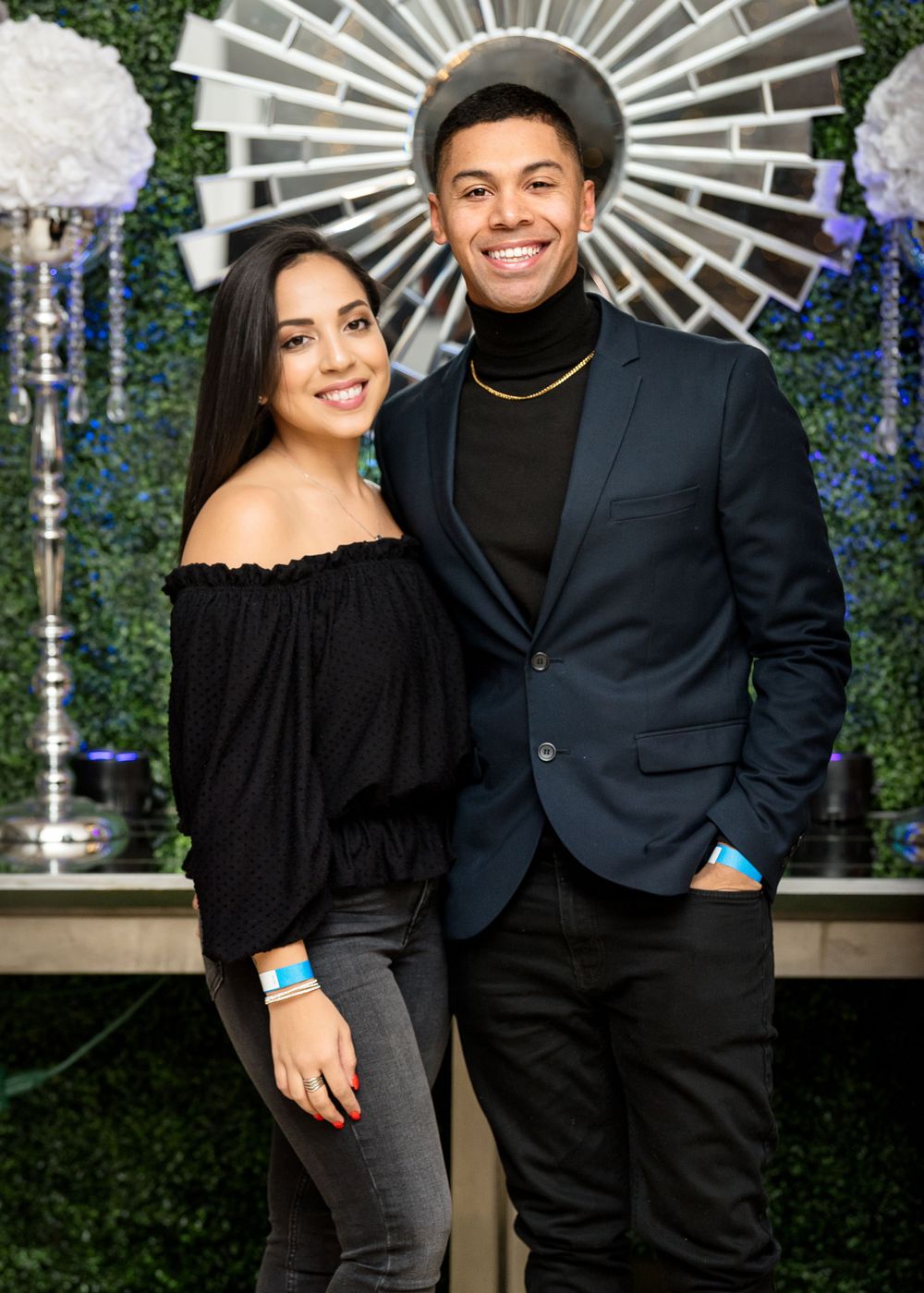 Couple in formal attire posing against decorative sunburst and floral wall backdrop.