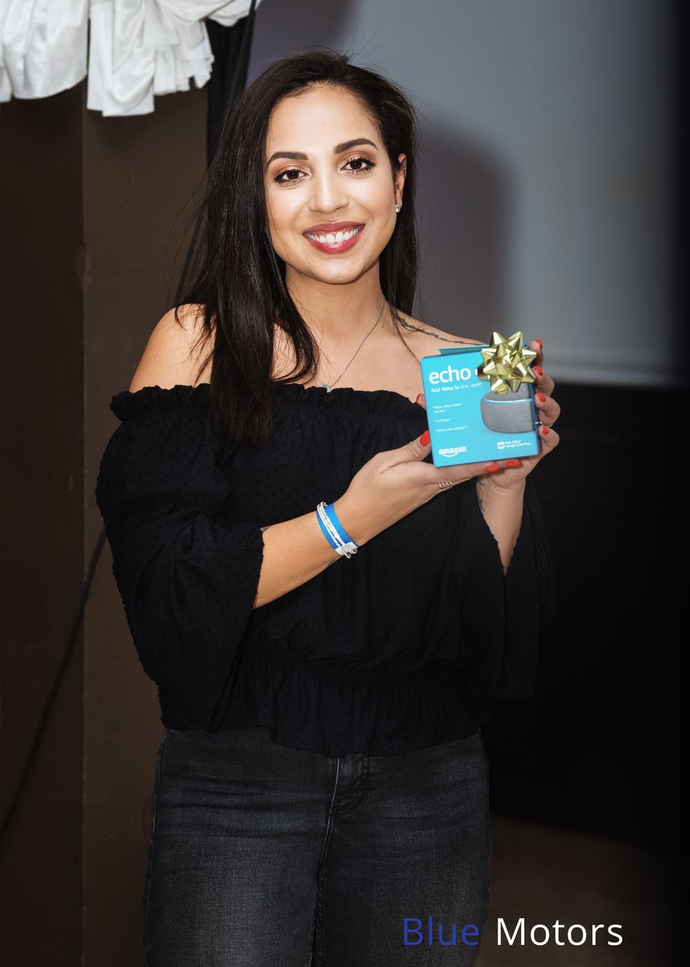 Woman in a black off-shoulder top holds up a small blue product package while smiling.