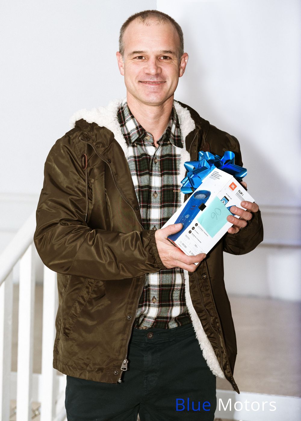 Person wearing brown jacket and plaid shirt holds blue-ribboned product package against white background.