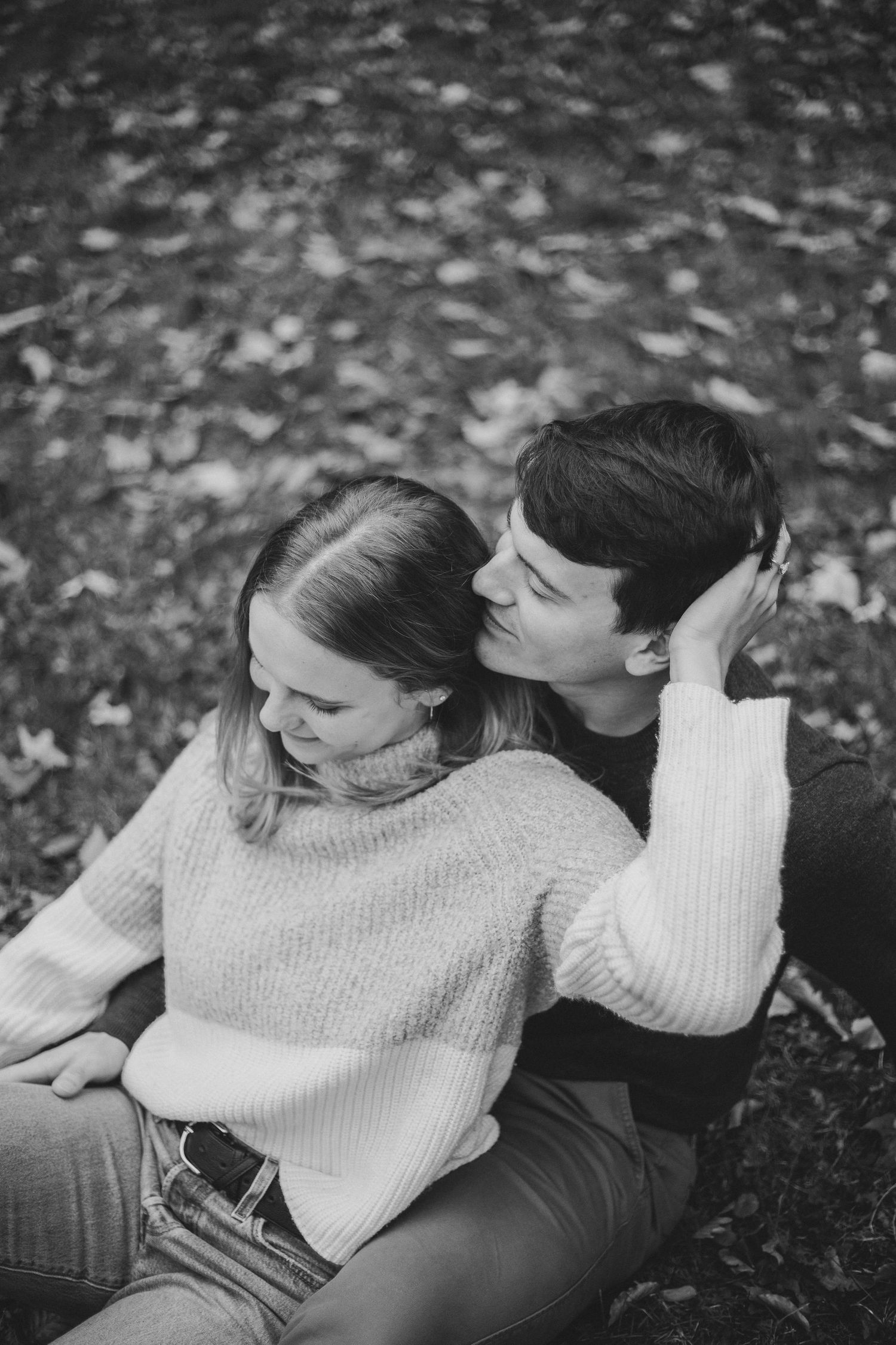 A romantic couple sits on fallen leaves sharing an intimate black and white moment.