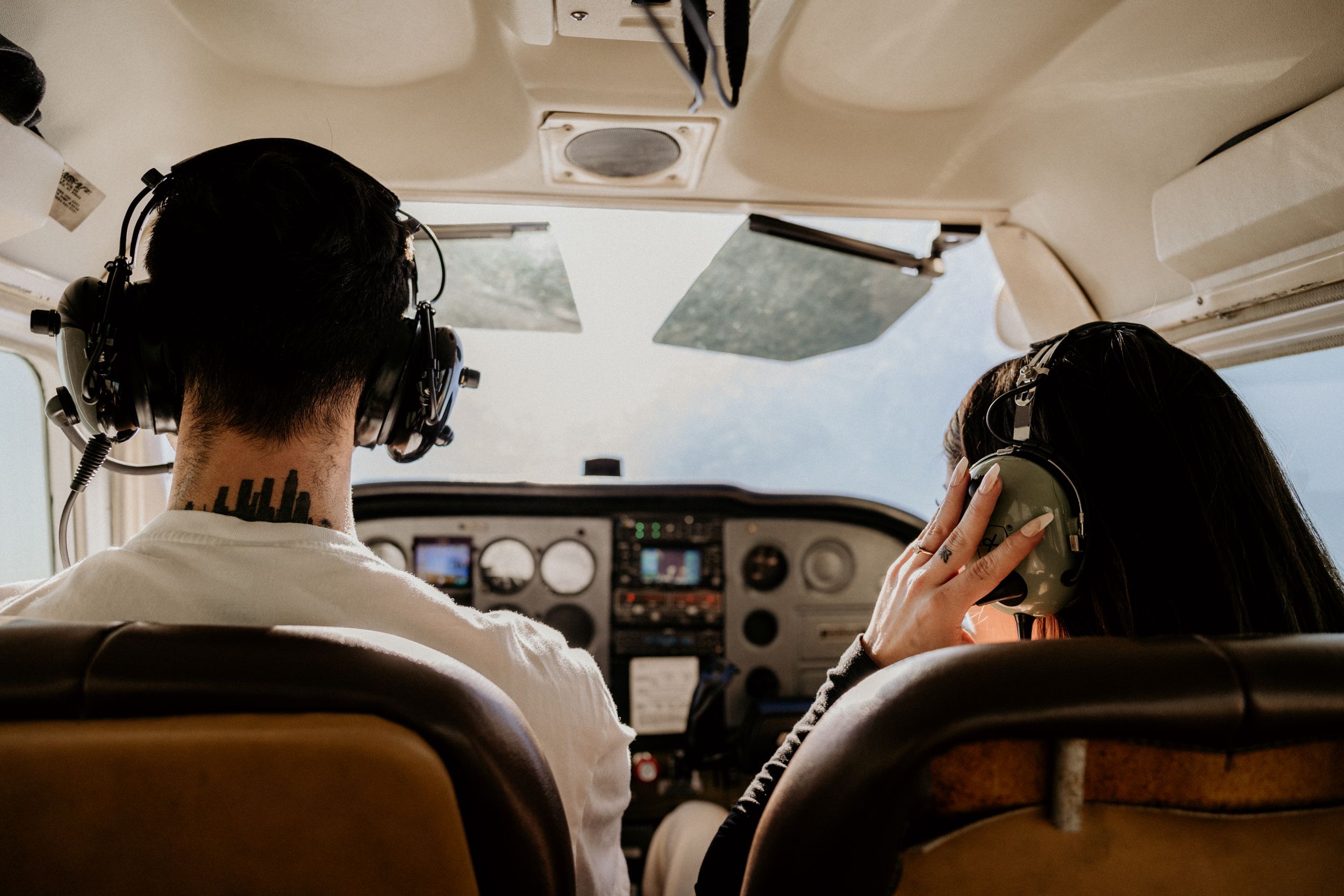 Man with neck tattoo and woman wearing headsets piloting small Cessna aircraft.