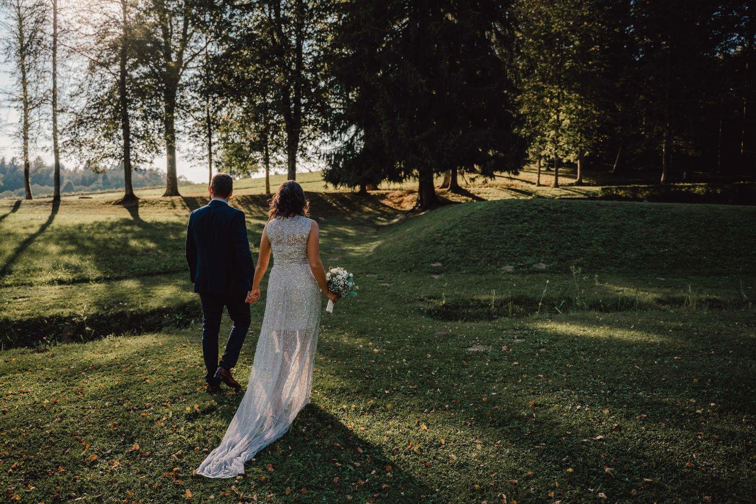 A couple walks through a sunlit park on their wedding day with trees casting long shadows on the grass.