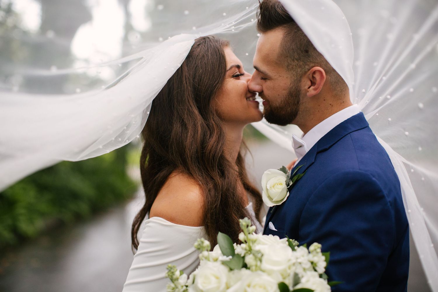Romantic wedding couple share an intimate moment under a flowing veil with white roses.