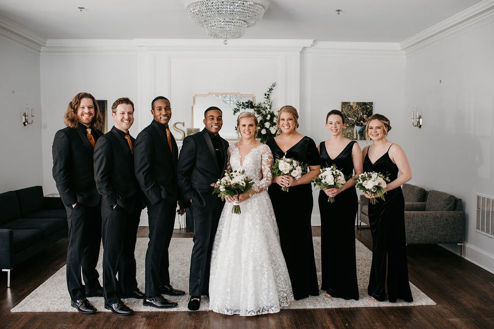 Bride and groom with wedding party dressed in black standing in an elegant white room.