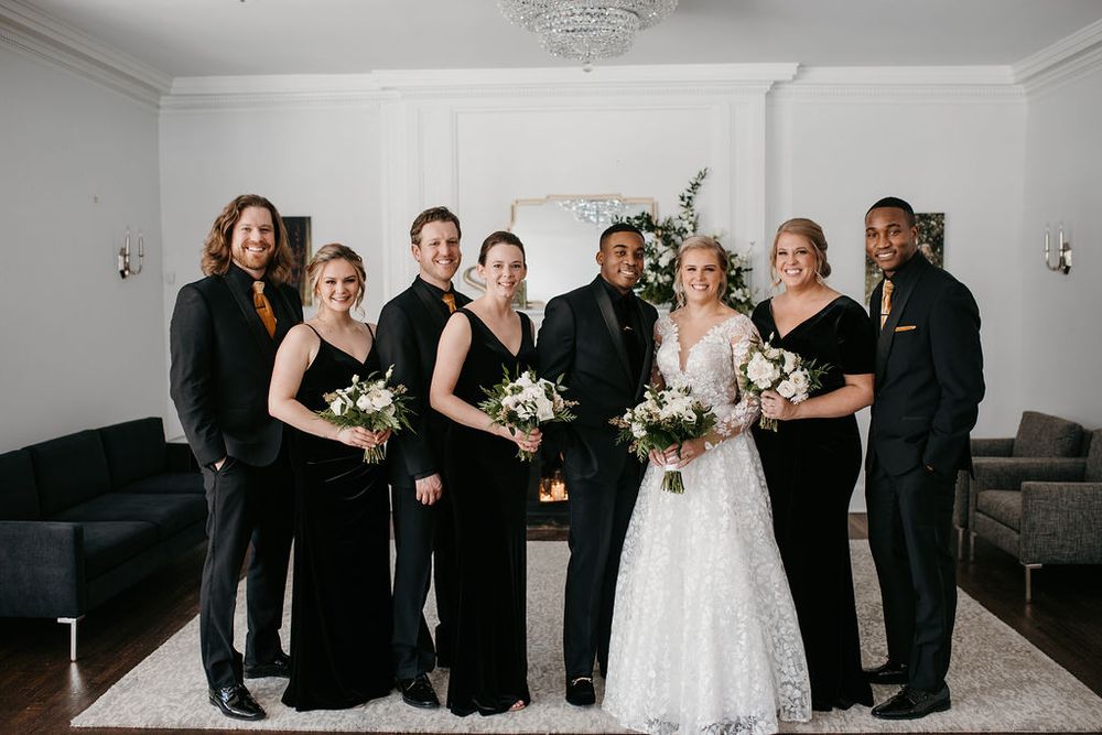 Wedding party in black attire posing with bride in lace gown near a fireplace.