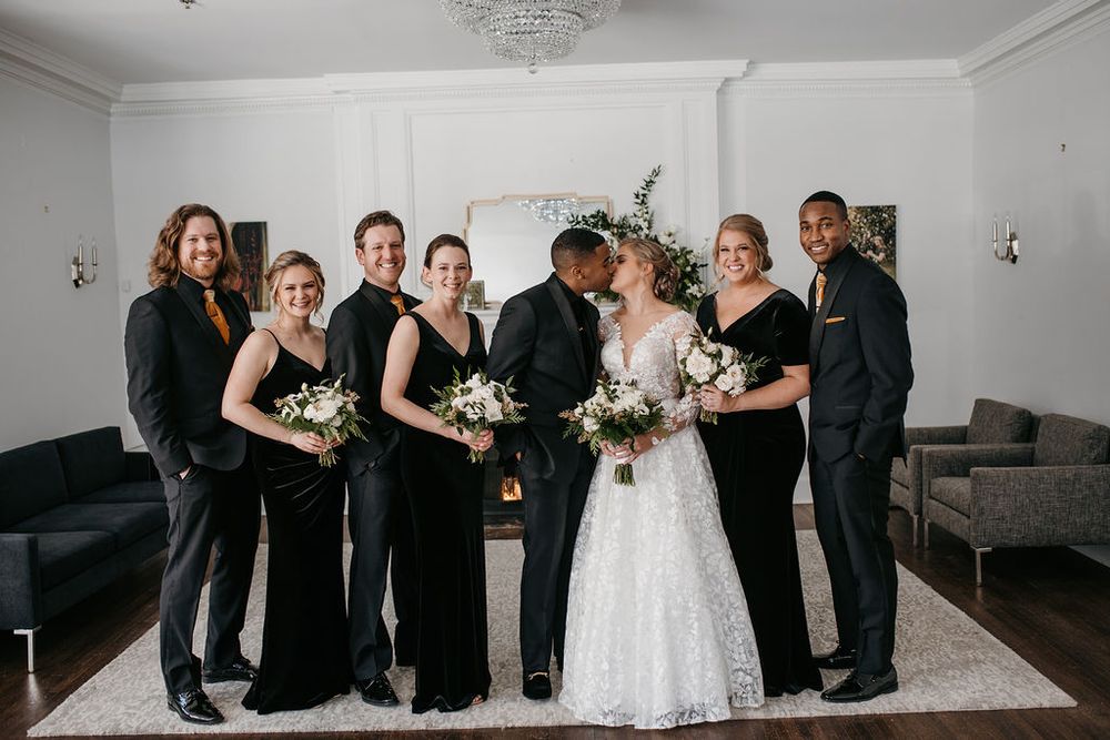 Bride and groom kissing surrounded by smiling wedding party dressed in black attire.