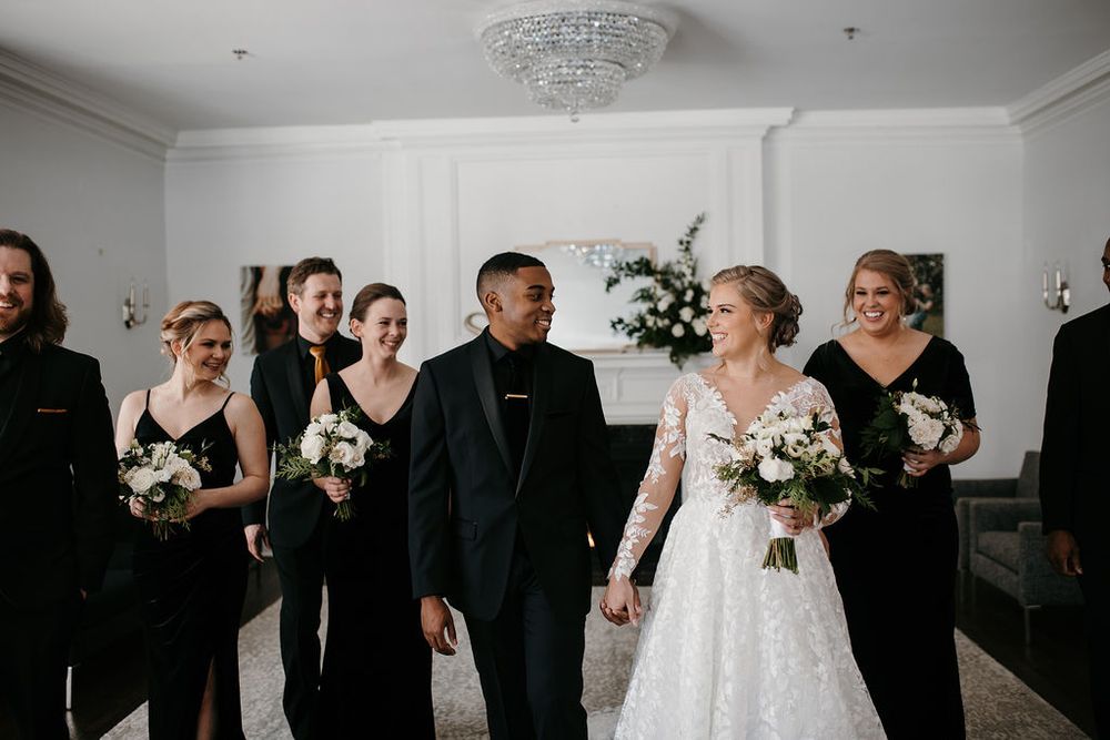 Bride in lace gown and groom in tuxedo walking with wedding party in black outfits.