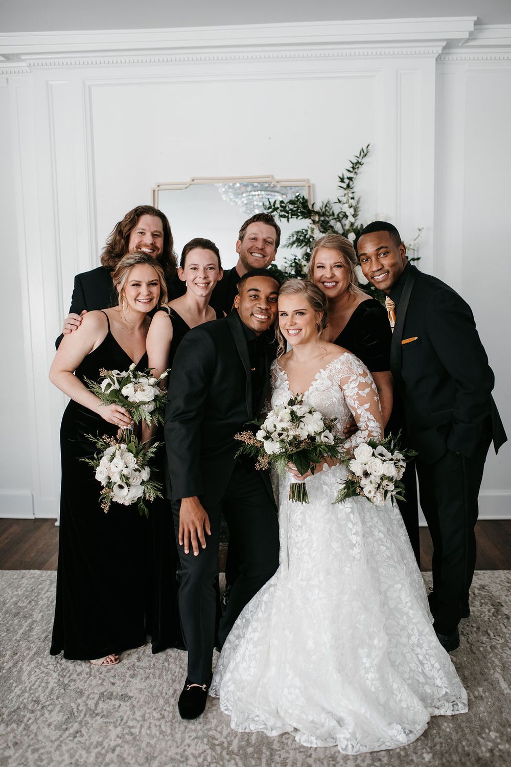 Wedding party group photo with bride holding white bouquet in front of a fireplace.