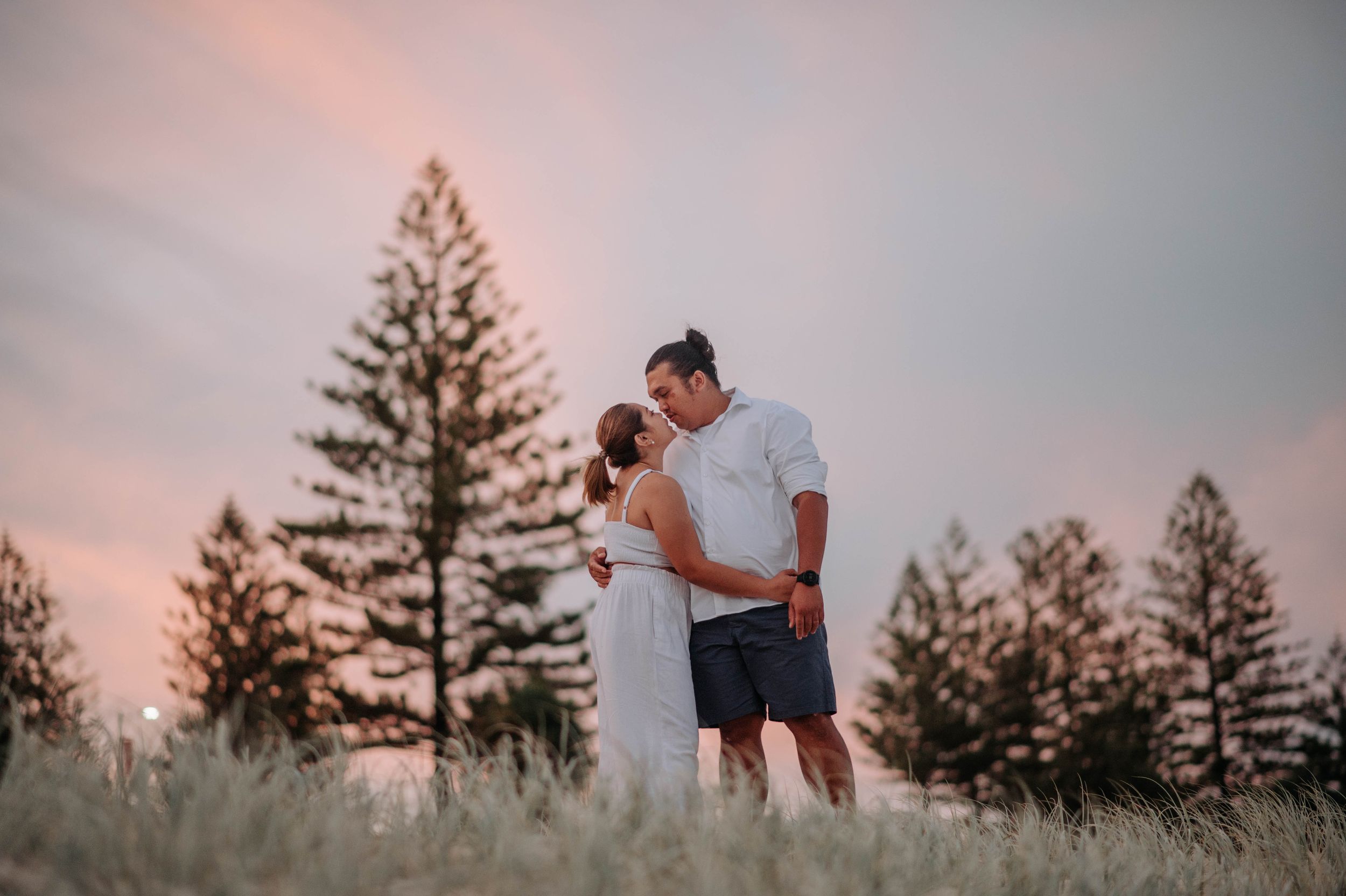 A romantic couple embraces in a grassy field at sunset with tall pine trees silhouetted in the background.
